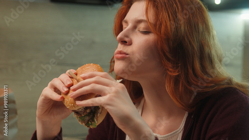 Obraz na plátně Young Caucasian woman female girl customer client eating junk fast food bite chewing burger hamburger cheeseburger eat pleasure diet cheat meal temptation indoors