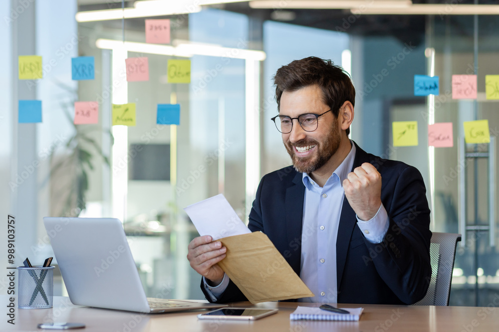 Businessman in suit happily reads successful notification from envelope at desk. Celebrates good news in contemporary office with notes and laptop. Concept of achievement, success, and communication. Businessman in suit happily reads successful notification from envelope at desk. Celebrates good news in contemporary office with notes and laptop. Concept of achievement, success, and communication.