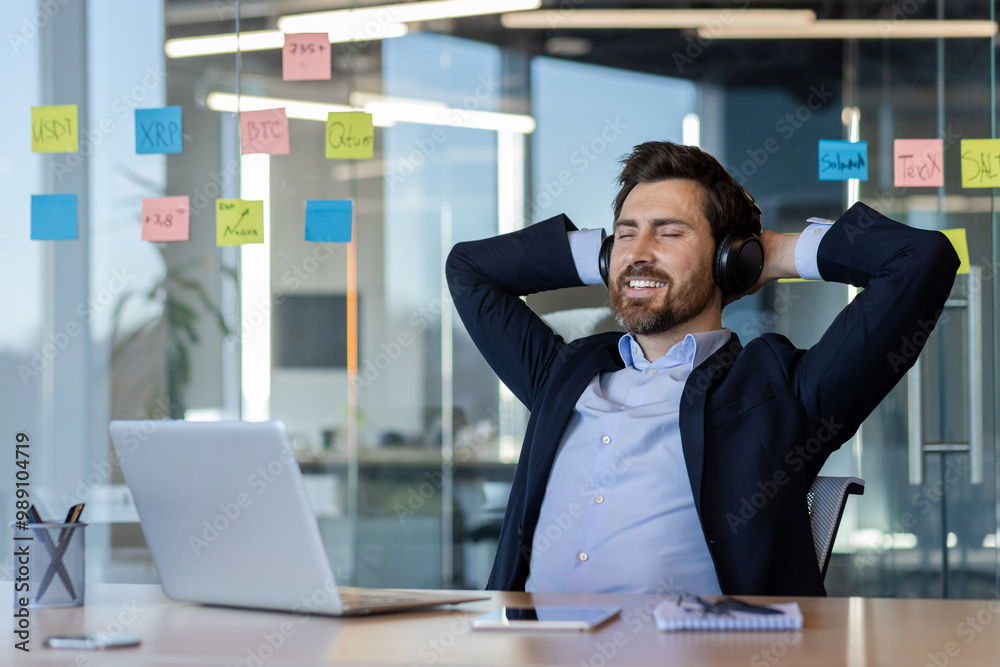 Business professional takes break in office, leaning back with headphones, smiling. Laptop on desk shows work environment. Sticky notes on glass wall indicate active projects. Business professional takes break in office, leaning back with headphones, smiling. Laptop on desk shows work environment. Sticky notes on glass wall indicate active projects.