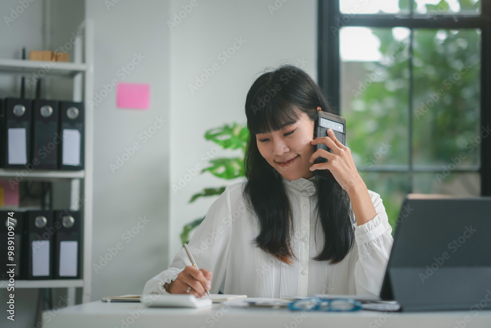 Asian accountant woman is smiling while talking on the phone and taking notes at her desk Asian accountant woman is smiling while talking on the phone and taking notes at her desk
