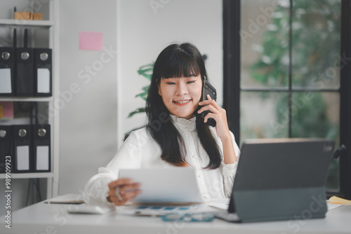 Asian businesswoman is smiling while talking on the phone and reading a document in her office