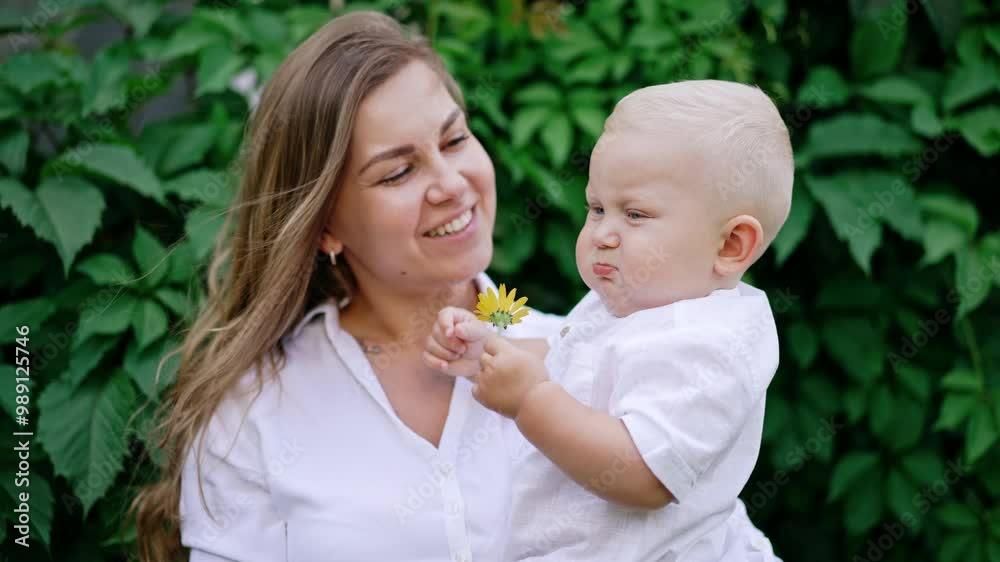 Smiling long-haired woman holding a little blond baby boy. Little kid holds a flower in hand and gives it to his mom to smell. Smiling long-haired woman holding a little blond baby boy. Little kid holds a flower in hand and gives it to his mom to smell.