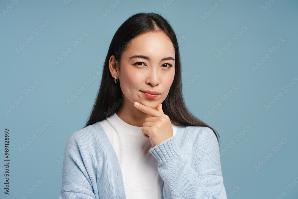 Pensive, young Asian woman touching chin and thinking looking at camera on blue background Pensive, young Asian woman touching chin and thinking looking at camera on blue background