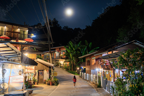 A woman walks down a street at night in Mae Kampong village with a full moon in the sky, Thailand