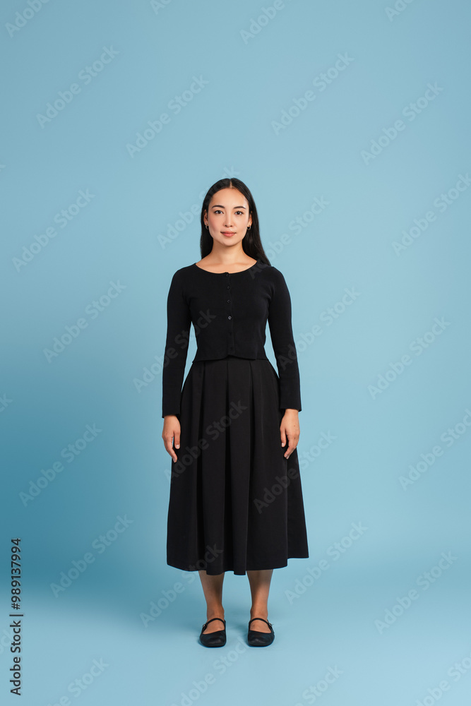 Beautiful Asian woman standing, looking at camera posing wearing black dress on blue background Beautiful Asian woman standing, looking at camera posing wearing black dress on blue background