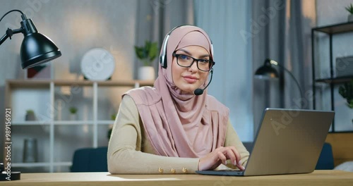 Woman in hijab with headset working on laptop at home and then looking into camera with happy face expression photo