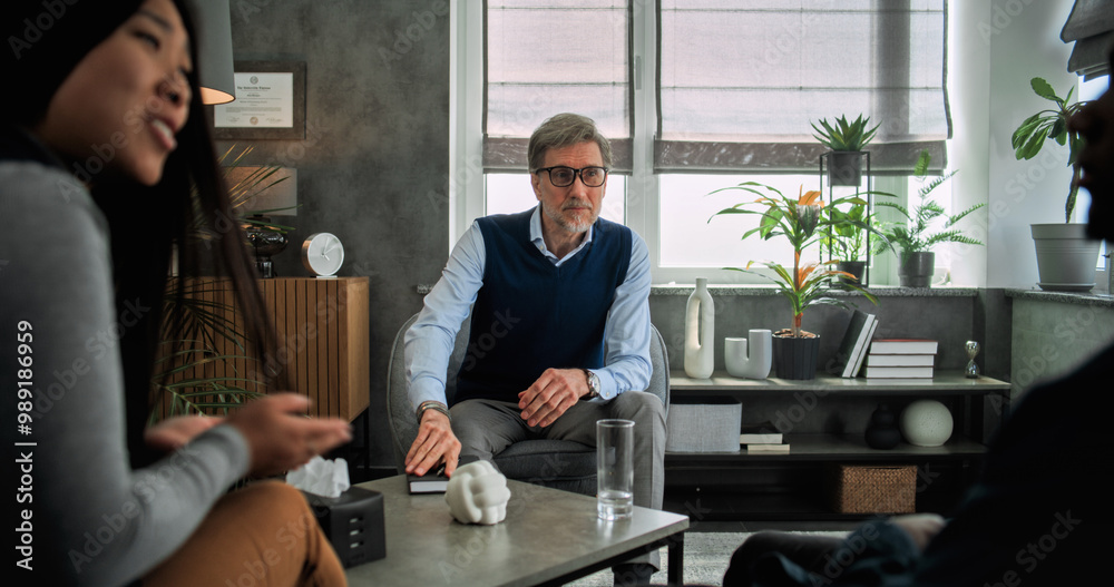 Back view of multiethnic couple sitting on chairs, discussing relationship problems, finding solution and compromise on couples therapy session. Mature therapist listens to young family, takes notes.