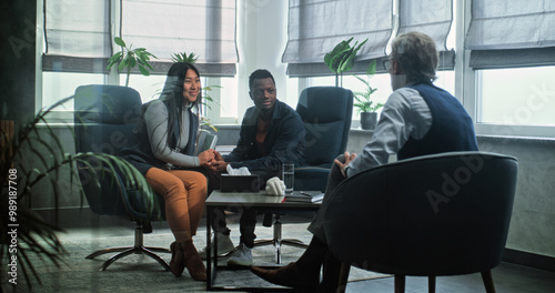 Multiethnic couple on counseling session with therapist. Mature psychologist talks to young family. African American man and Asian woman sit on chairs holding hands after quarrel on couples therapy.