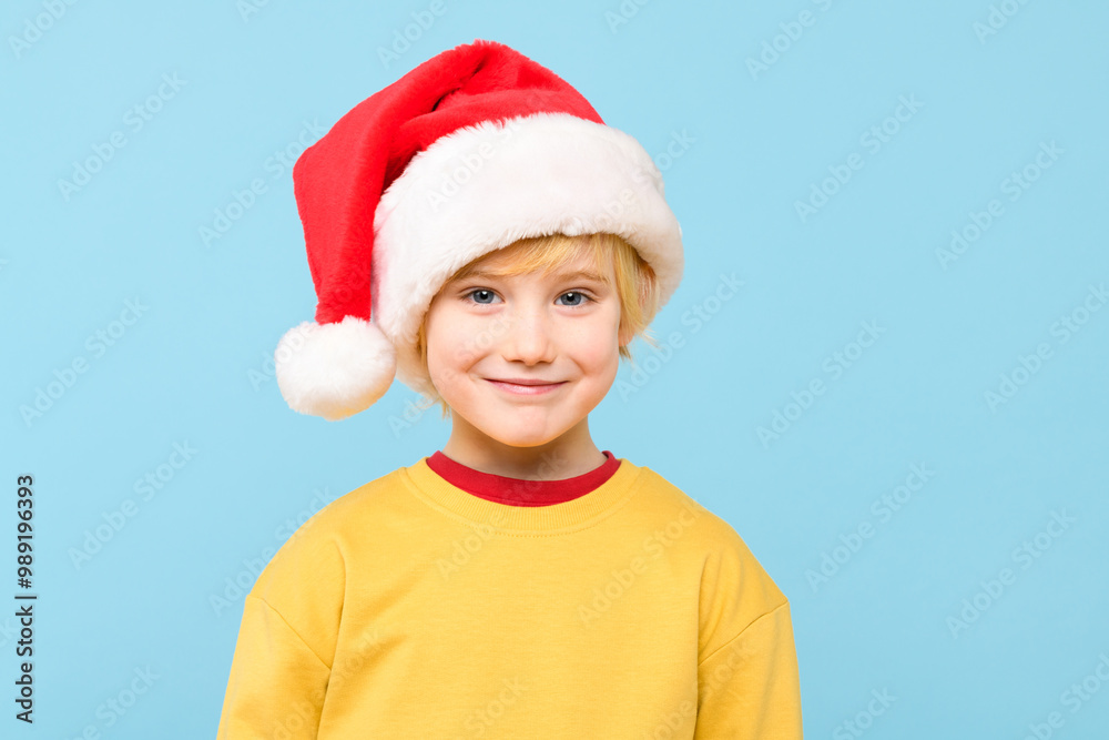 Cute young caucasian preschooler boy wearing santa hat, smiling while looking at camera. Studio portrait isolated over pastel blue background. Cute young caucasian preschooler boy wearing santa hat, smiling while looking at camera. Studio portrait isolated over pastel blue background.