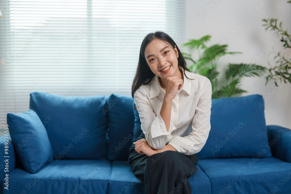 Young asian businesswoman is smiling while sitting on a sofa at home, with her hand on her chin Young asian businesswoman is smiling while sitting on a sofa at home, with her hand on her chin
