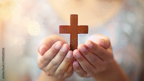 Both hands hold a small wooden cross against a soft, light colored background. Prayer and personal devotion make it perfect for topics related to faith, Christian worship.