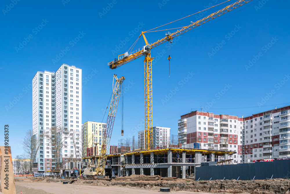 A construction site where tower and crawler cranes perform lifting work. A new house is being built on the site of the old houses. A road is being paved near the house under construction. A construction site where tower and crawler cranes perform lifting work. A new house is being built on the site of the old houses. A road is being paved near the house under construction.