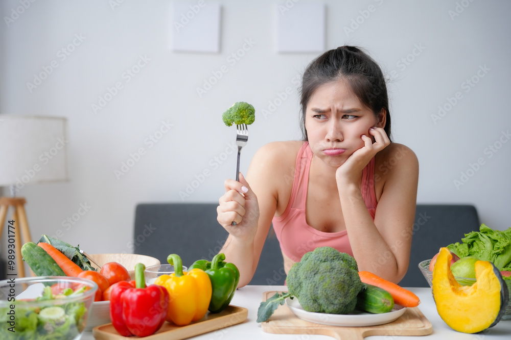 Young sporty woman holding broccoli with bored expression while being on diet with many kinds of vegetables on table in living room Young sporty woman holding broccoli with bored expression while being on diet with many kinds of vegetables on table in living room