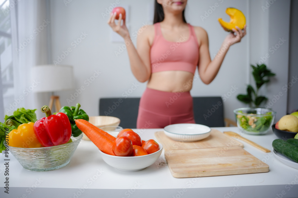 Young woman in sportswear is holding fruits, standing behind a kitchen table full of fresh vegetables Young woman in sportswear is holding fruits, standing behind a kitchen table full of fresh vegetables