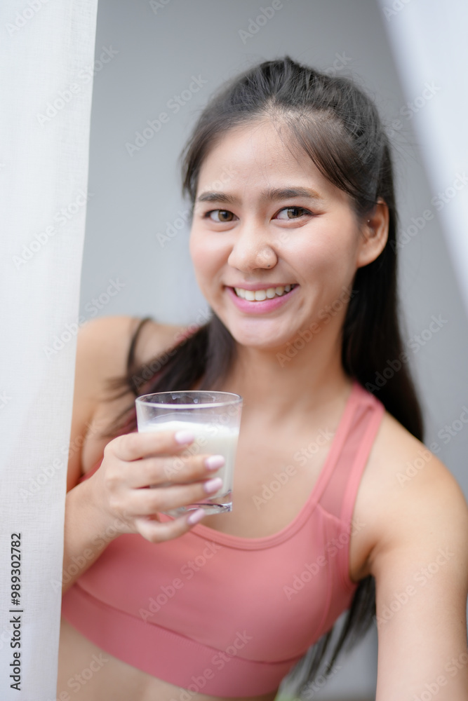 Asian woman smiles and drinks milk after exercising at home, radiating freshness and vitality. She promotes a healthy lifestyle, emphasizing wellness and nutrition Asian woman smiles and drinks milk after exercising at home, radiating freshness and vitality. She promotes a healthy lifestyle, emphasizing wellness and nutrition