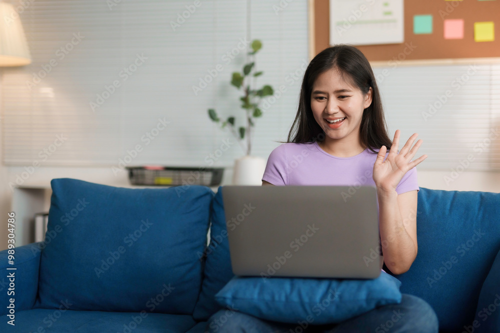 Young woman is sitting on a blue couch in her living room, waving to someone on her laptop during a video call Young woman is sitting on a blue couch in her living room, waving to someone on her laptop during a video call