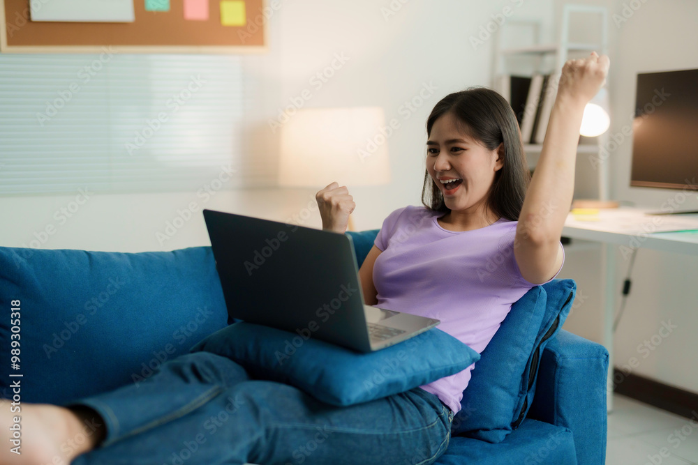 Naklejka premium Young asian freelancer is raising her arms in celebration while working on her laptop, expressing joy and excitement in her home office