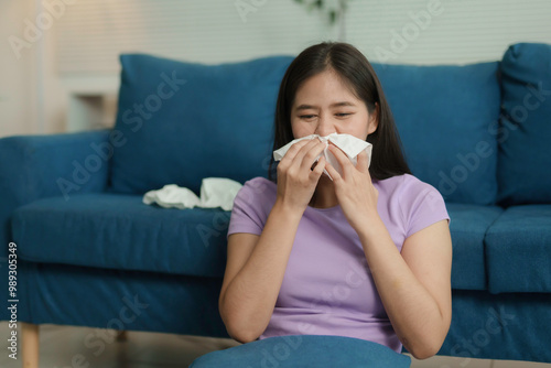 Young woman sitting on the floor in the living room blowing her nose, suffering from a cold or flu virus