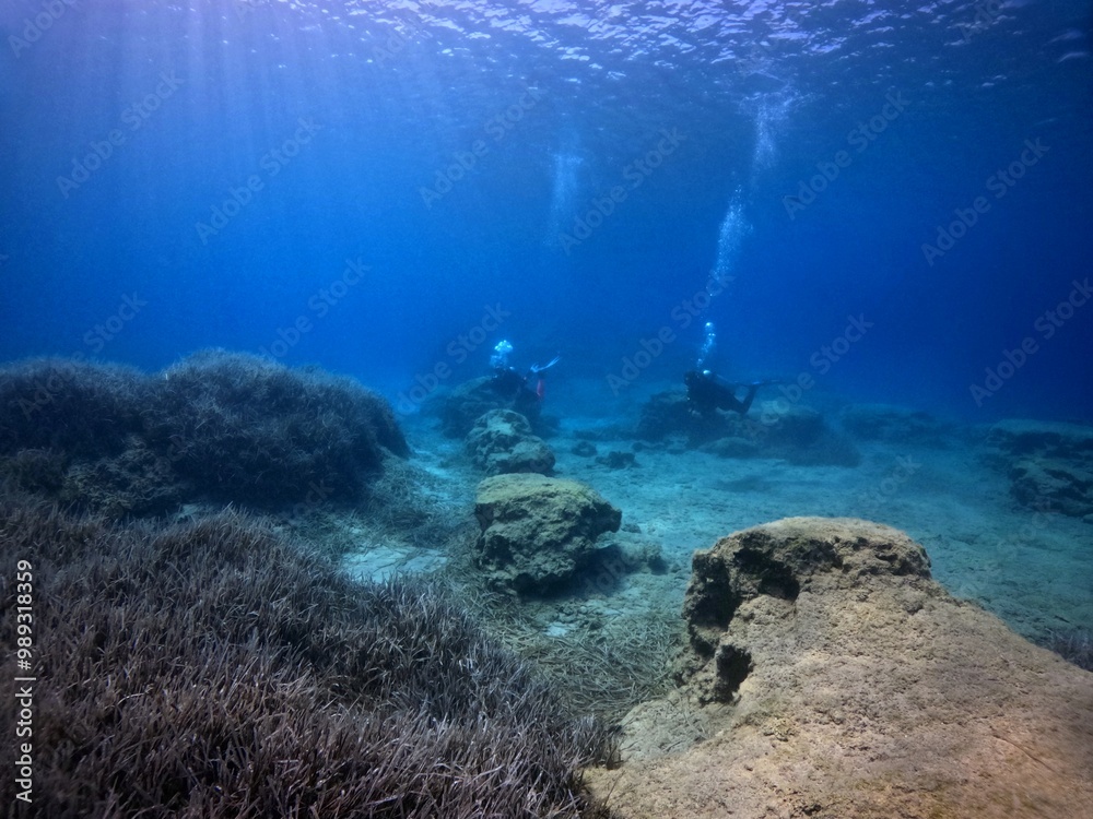 Obraz premium Rocky underwater landscape with pair of swimming scuba divers. Underwater photography from scuba diving exploration trip. Divers in the ocean, travel photo. Bubbles from breathing in the water.