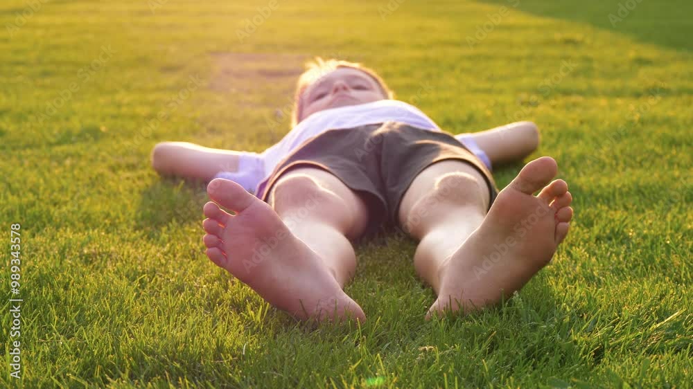 The child is lying on the green grass. The child is swinging their bare feet. The child is enjoying nature and relaxing outdoors. The concept is a happy childhood and a family picnic. The child is lying on the green grass. The child is swinging their bare feet. The child is enjoying nature and relaxing outdoors. The concept is a happy childhood and a family picnic.