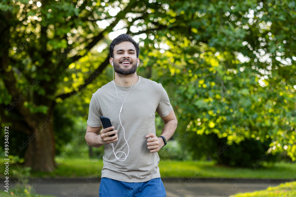 Smiling Indian man in headphones and phone running in the park and doing sports Smiling Indian man in headphones and phone running in the park and doing sports