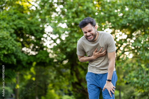 Young Indian man doing sports and jogging in the park, looking forward, holding hand to chest and grimacing from severe heart pain