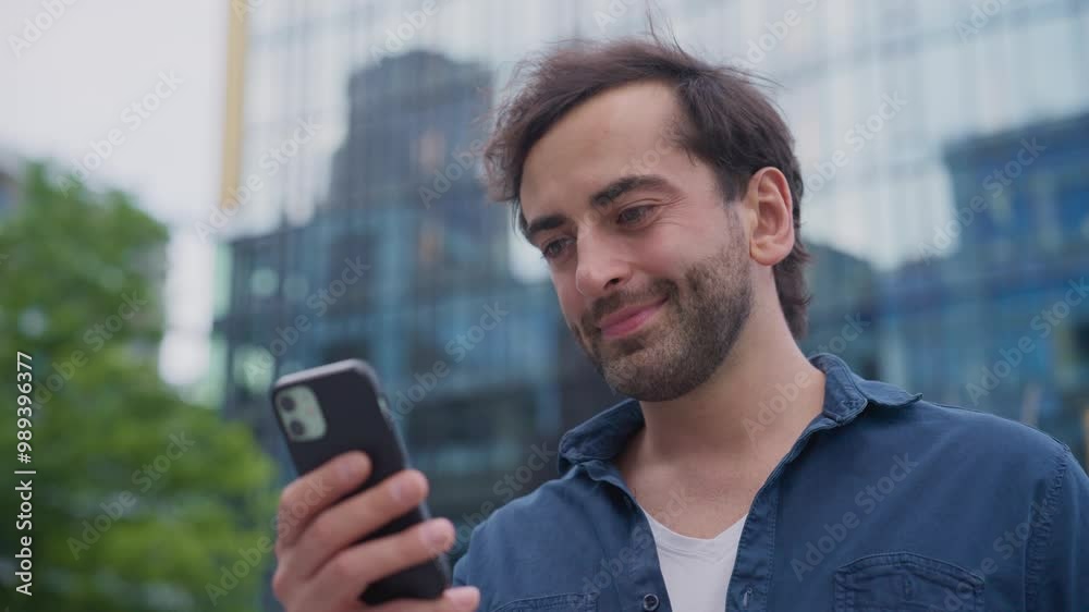Positive Caucasian man cheerfully smiling while reading message on smartphone. Bearded male using phone for online communication. Messaging with someone in chat. Modern lifestyle. City center. Positive Caucasian man cheerfully smiling while reading message on smartphone. Bearded male using phone for online communication. Messaging with someone in chat. Modern lifestyle. City center.