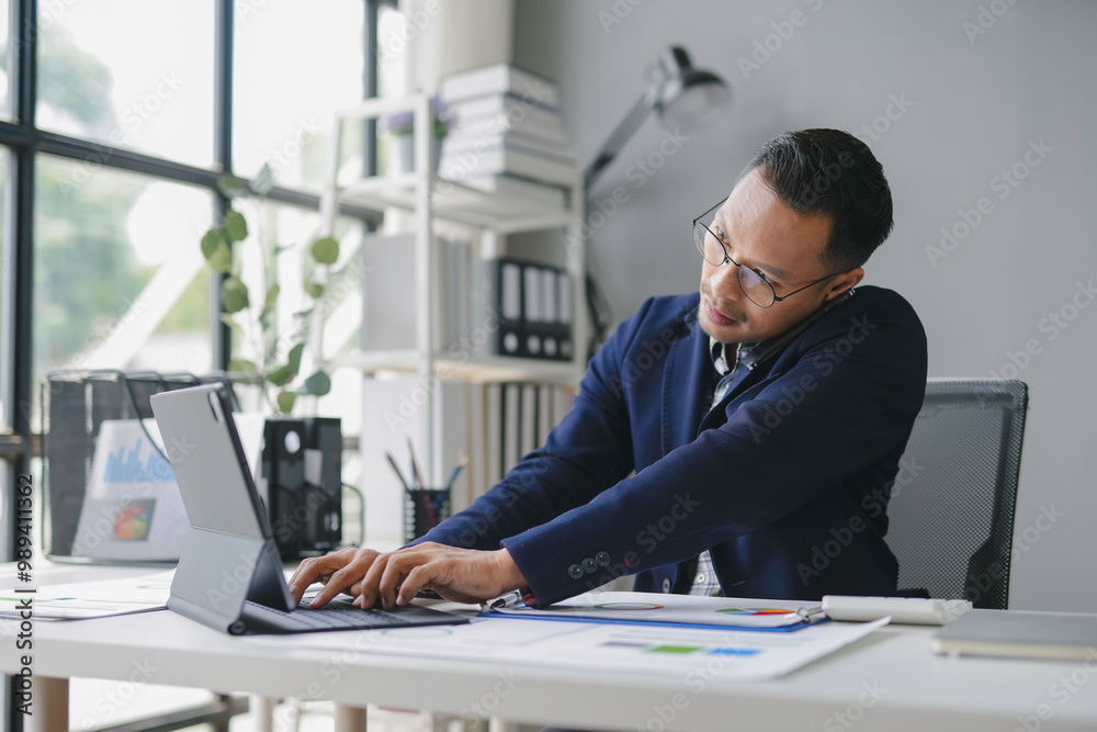 Young businessman is working on a tablet computer at his desk in a modern office, surrounded by paperwork and digital devices Young businessman is working on a tablet computer at his desk in a modern office, surrounded by paperwork and digital devices