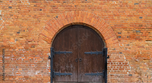 Fotografie Old brick wall with arch and closed wooden gate with old-style door hinges and r