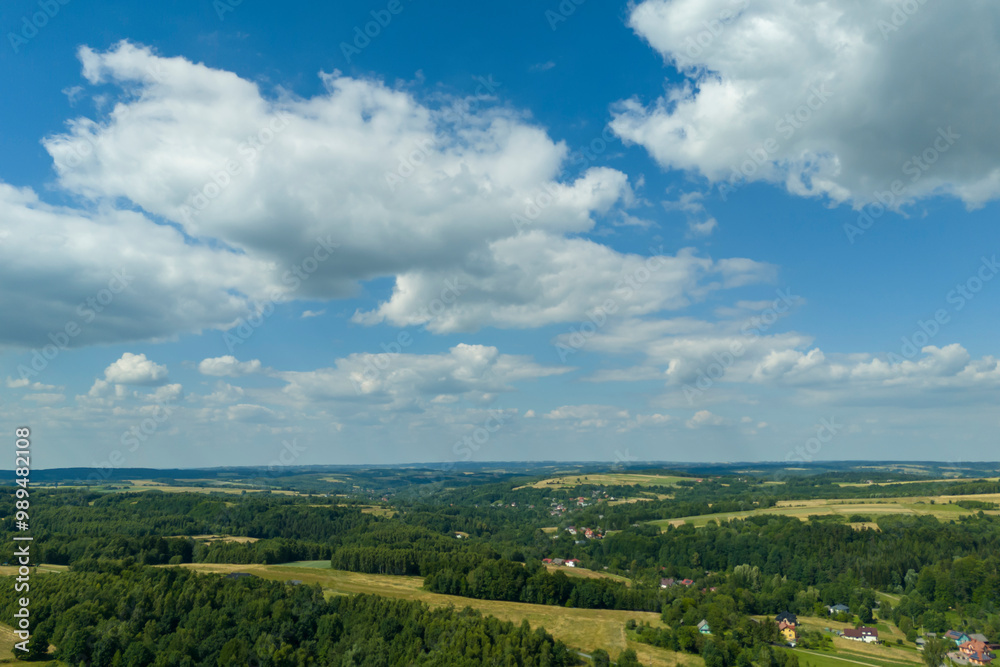 Obraz premium A stunning aerial view of a serene landscape featuring green fields and trees under a bright blue sky adorned with fluffy clouds.