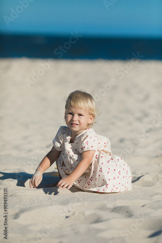 2 years old girl in white clothes sits on the white sand on the beach in summer