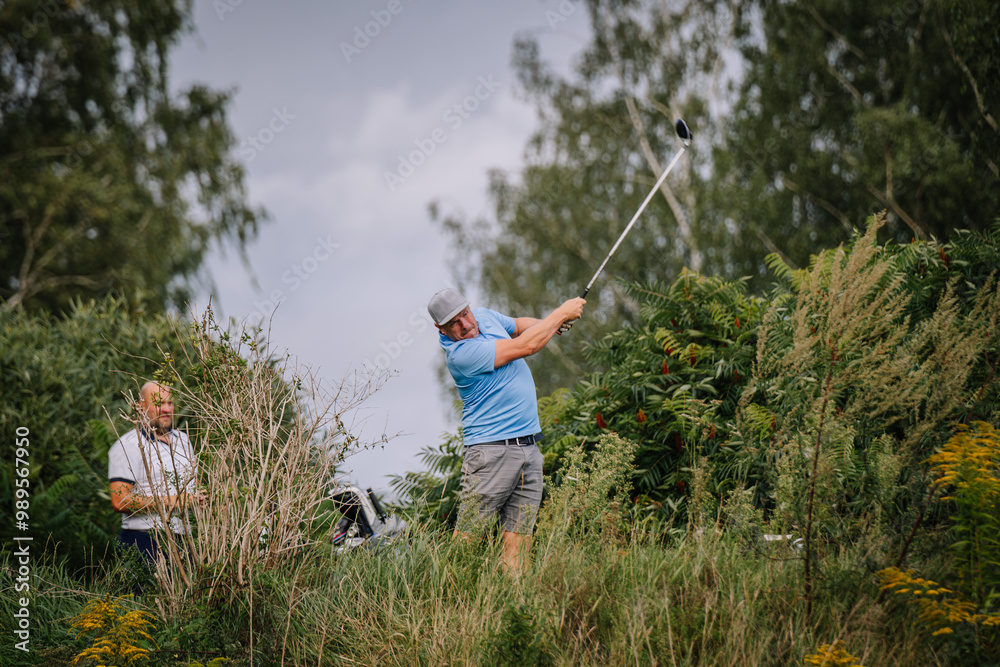 A golfer swings his club in a lush outdoor setting, surrounded by greenery, while another man observes in the background. A golfer swings his club in a lush outdoor setting, surrounded by greenery, while another man observes in the background.