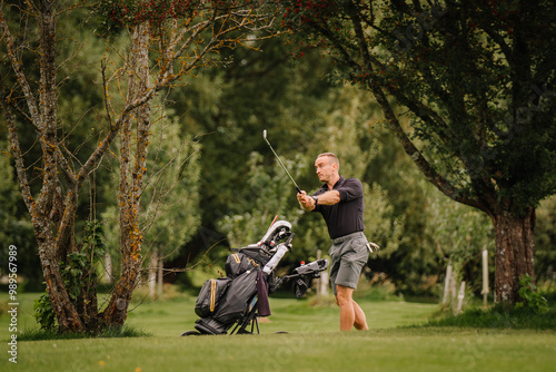 A golfer in mid-swing near a tree on the golf course, with his golf cart and bag beside him, surrounded by lush greenery.