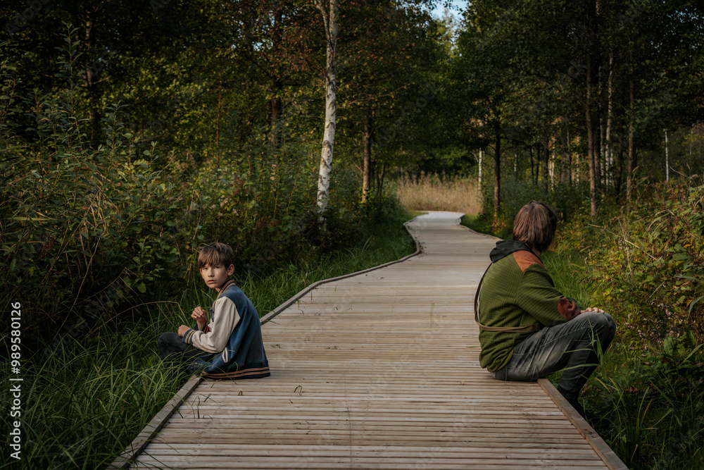 Fototapeta premium A young boy and an older teenager sit on a wooden pathway surrounded by greenery. The younger boy holds a grass blade, while the teenager looks thoughtful, gazing into the distance.