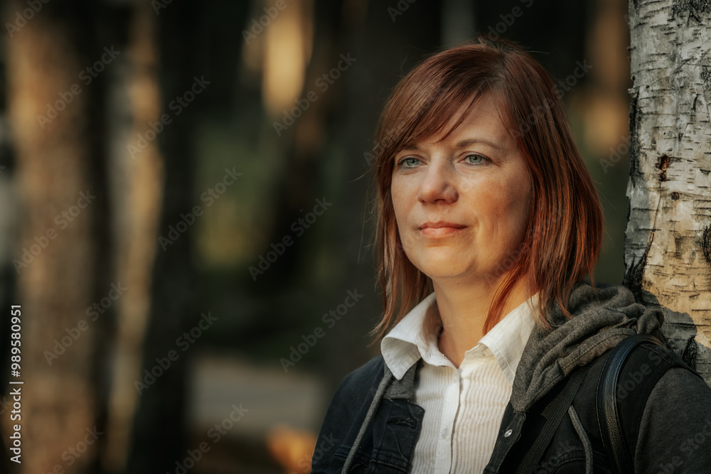 A thoughtful woman with red hair stands leaning against a birch tree in a forest. The soft glow of sunset illuminates her face, creating a peaceful and contemplative atmosphere. A thoughtful woman with red hair stands leaning against a birch tree in a forest. The soft glow of sunset illuminates her face, creating a peaceful and contemplative atmosphere.