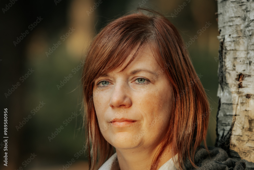 A thoughtful woman with red hair stands leaning against a birch tree in a forest. The soft glow of sunset illuminates her face, creating a peaceful and contemplative atmosphere. A thoughtful woman with red hair stands leaning against a birch tree in a forest. The soft glow of sunset illuminates her face, creating a peaceful and contemplative atmosphere.
