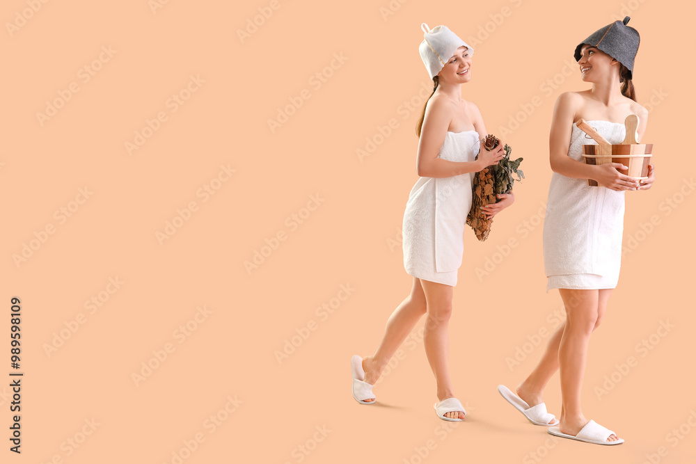 Young women in sauna hats with whisk and bucket of water on beige background Young women in sauna hats with whisk and bucket of water on beige background