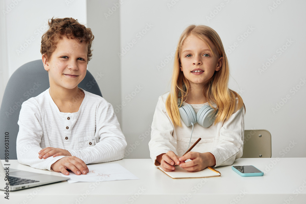 Happy children studying together online at home boy and girl sitting at a table in the living room, using laptops and headphones for elearning They are engaged, concentrated, and enjoying their online