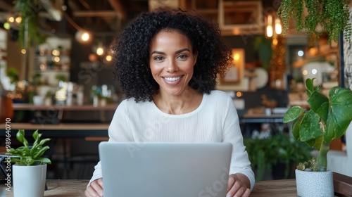 A cheerful woman with curly hair works on her laptop in a warmly lit cafe, surrounded by green plants and warm ambiance, suggesting productivity and relaxation.