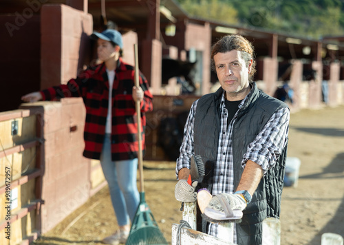 Adult male stables worker in plaid shirt holding brushes and gloves before cleaning horses from dirt in ranch photo