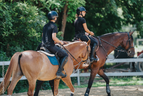 Two individuals riding horses in an outdoor equestrian arena with lush greenery in the background. photo