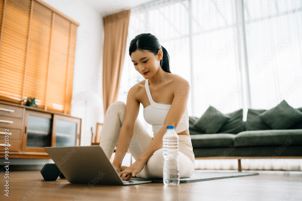 Fototapeta premium Young Asian woman sitting on the floor on yoga mat in front of laptop and training at home. Beautiful smiling woman doing stretching exercise while communicates with a trainer online before workout