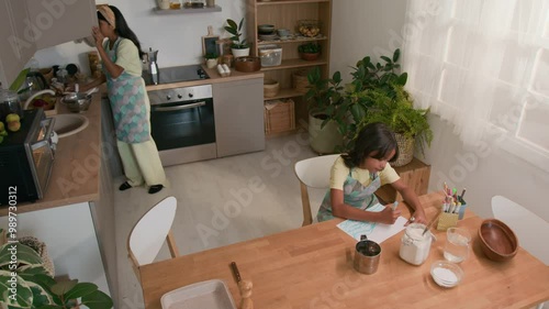 Top view of Hispanic mother in apron and hairband bringing utensils and ingredients from cupboard to table, preparing to bake cookies with little daughter who drawing in kitchen photo