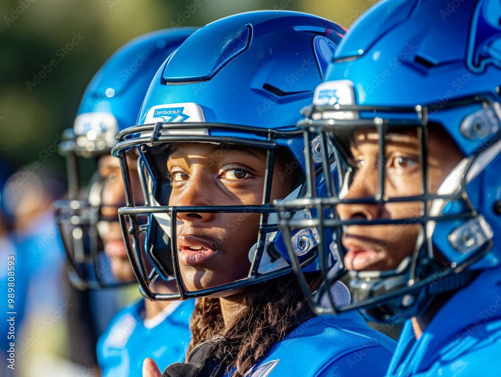 Three football players wearing blue helmets stand together. One of them has a black face mask on. The other two players are looking at the camera