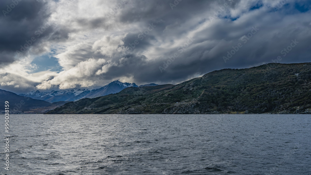 The picturesque coastline. Snow-capped mountain peaks of the Andes against a blue sky, clouds. Green vegetation on the coastal hills. The city buildings of Ushuaia on the shore. Ripples on the ocean