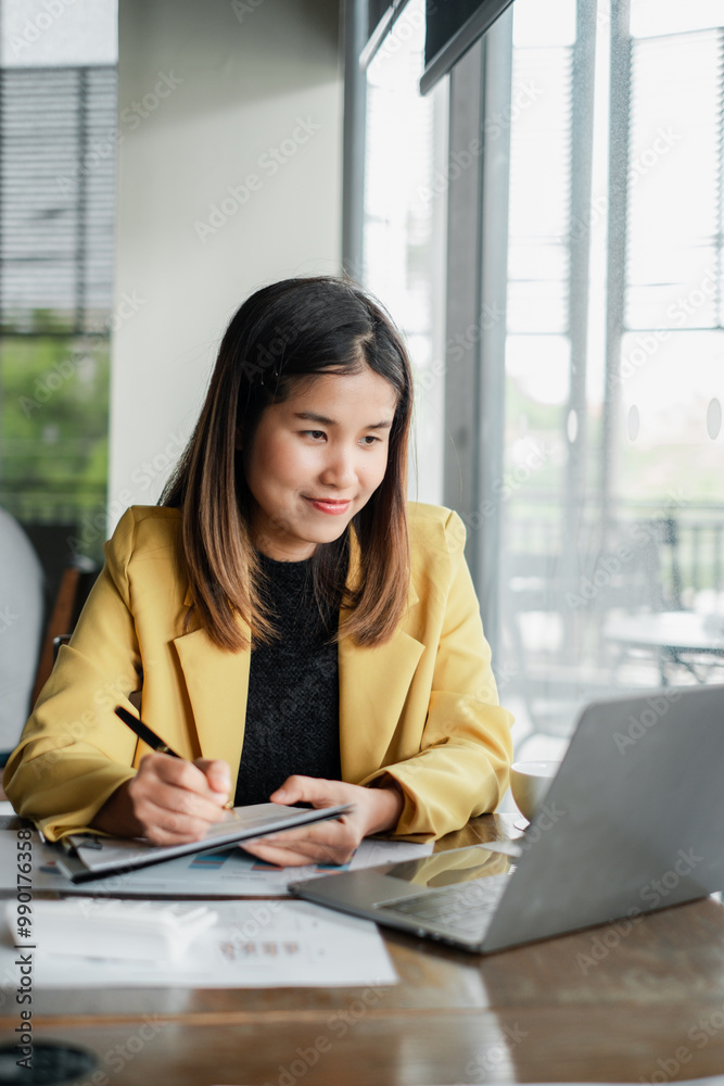 A young woman in a yellow blazer working on a laptop in a bright, modern office space with large windows and natural light. A young woman in a yellow blazer working on a laptop in a bright, modern office space with large windows and natural light.
