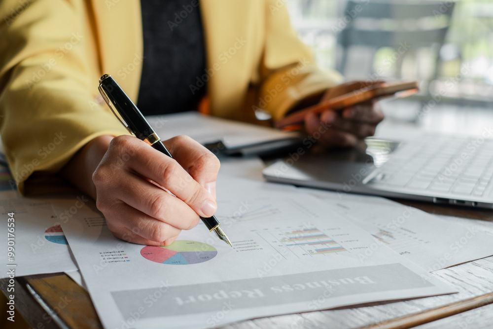 Close-up of a business professional analyzing financial reports with a pen and smartphone at an office desk. Close-up of a business professional analyzing financial reports with a pen and smartphone at an office desk.