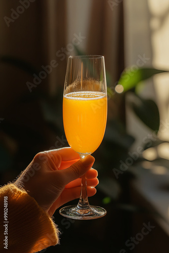 hand holding flute glass of mimosa, illuminated by soft sunlight, creates warm and inviting atmosphere. vibrant orange drink contrasts beautifully with greenery in background