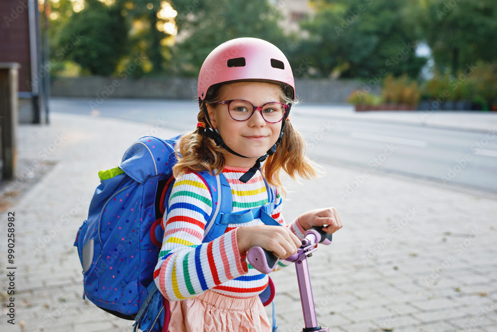 Little happy girl on way to school by pushing scooter. Elementary school child riding in the city, with big satchel. Kid with pink helmet. Safe route to school and movement for children concept. Little happy girl on way to school by pushing scooter. Elementary school child riding in the city, with big satchel. Kid with pink helmet. Safe route to school and movement for children concept.