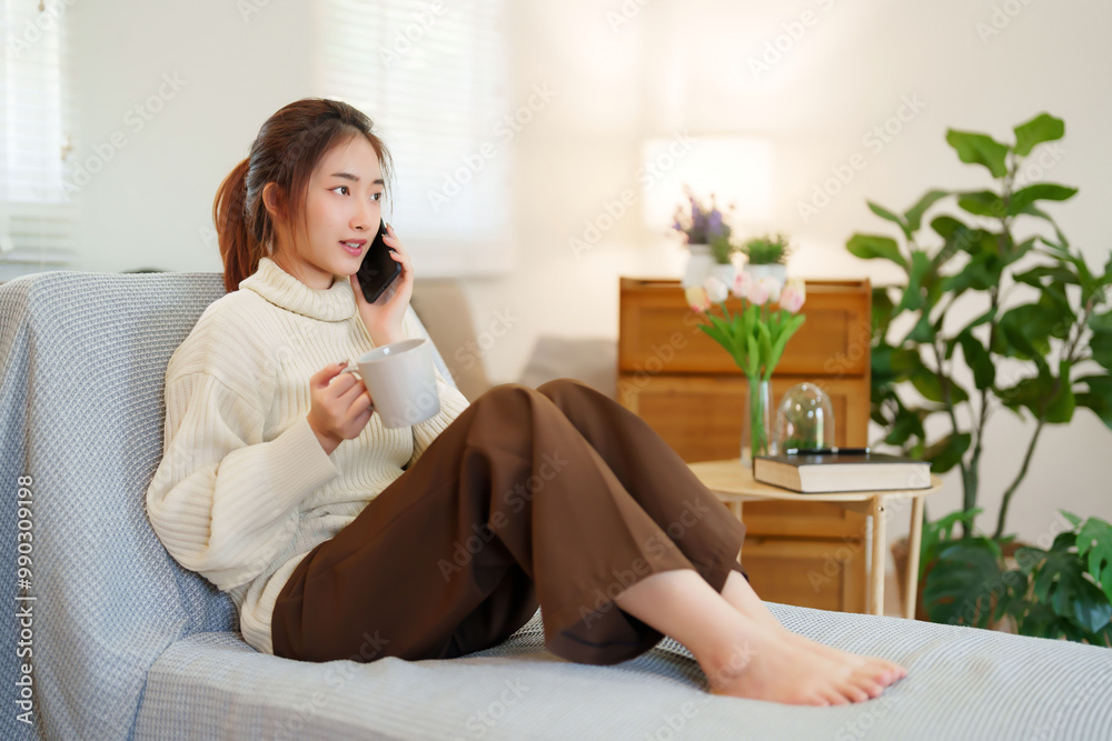 Young asian women in sweater talk to calling on smartphone and drinking coffee while sitting on comfortable couch to relaxation and doing activity for spending time with slow life lifestyle at home Young asian women in sweater talk to calling on smartphone and drinking coffee while sitting on comfortable couch to relaxation and doing activity for spending time with slow life lifestyle at home